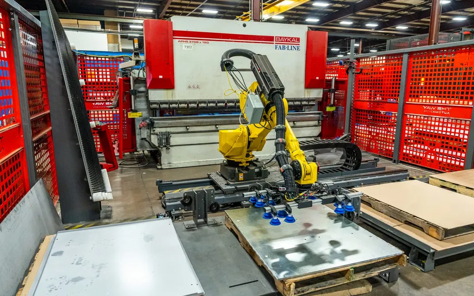 A FANUC robot on a seventh-axis rail with a blue vacuum-cup gripper lifting a galvanized sheet from a pallet, in front of a Baykal press brake inside red fencing