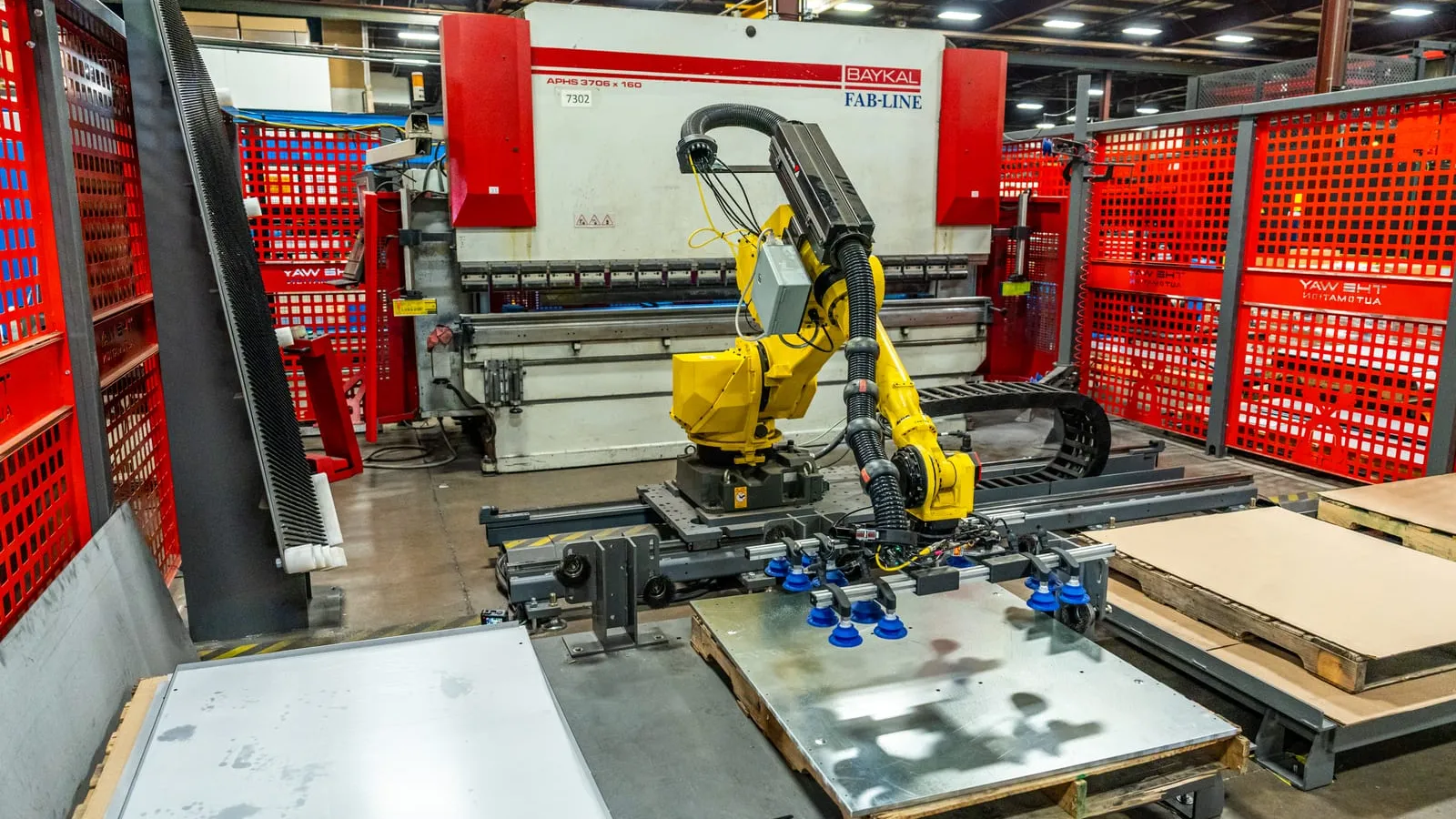 A FANUC robot on a seventh-axis rail with a blue vacuum-cup gripper lifting a galvanized sheet from a pallet, in front of a Baykal press brake inside red fencing