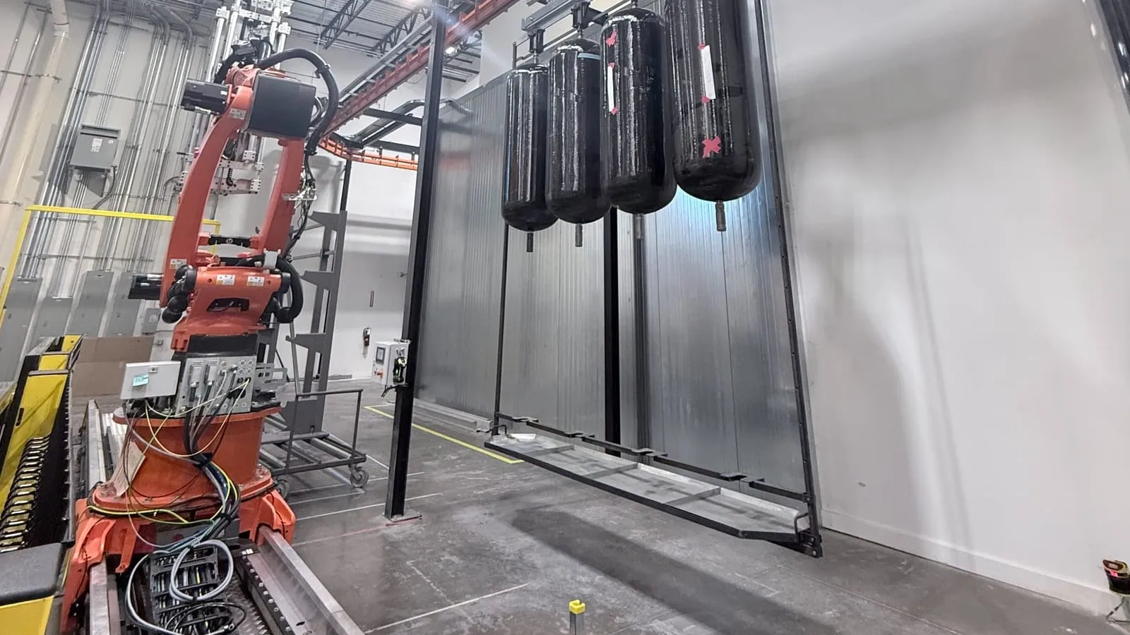 A six-axis robot on a long linear rail beside a curing oven, with four black composite pressure vessels hanging on a rack