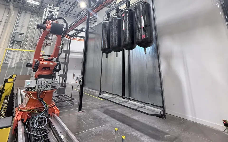 A six-axis robot on a long linear rail beside a curing oven, with four black composite pressure vessels hanging on a rack