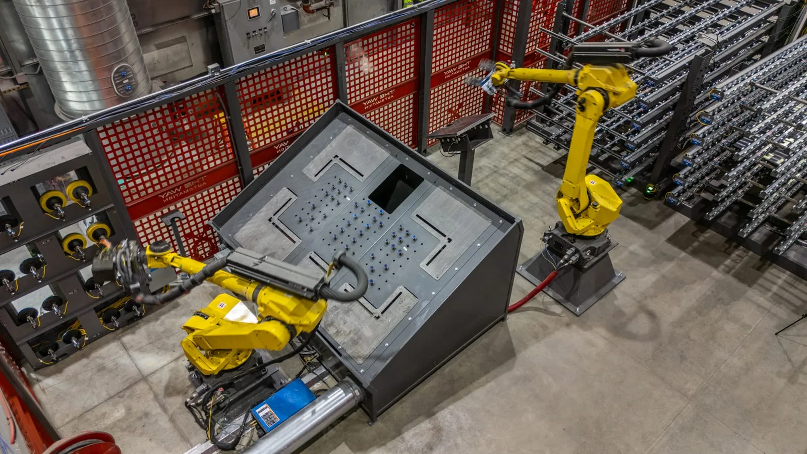 Two FANUC robots handling a large formed sheet-metal panel at the fixture table in the sanding cell