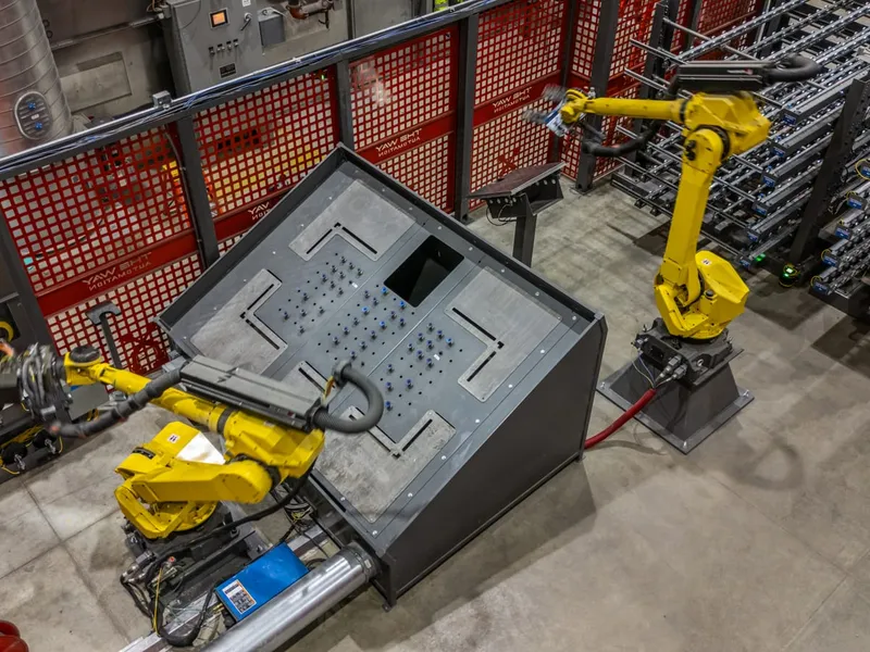 Two FANUC robots handle a large formed enclosure panel inside a fenced finishing cell