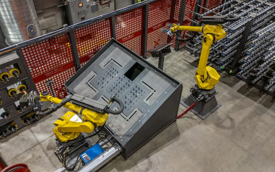 Two FANUC robots handling a large formed sheet-metal panel at the fixture table in the sanding cell