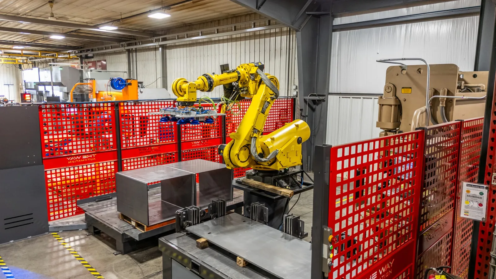 A yellow FANUC robot with a vacuum gripper handles sheet metal blanks inside a red guarded cell beside a press brake