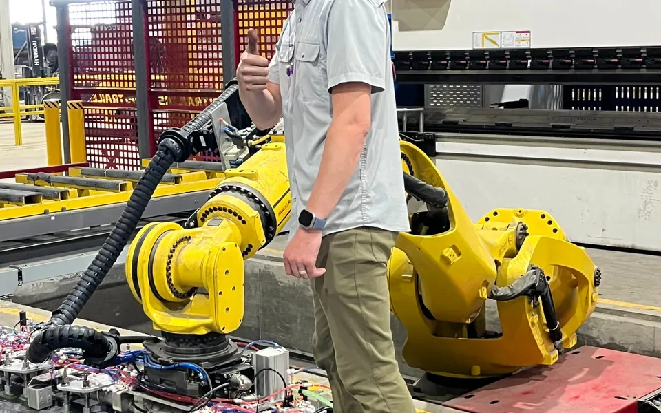 A technician beside a FANUC robot with a long vacuum-cup bar in front of a press brake, on the heavy tandem project