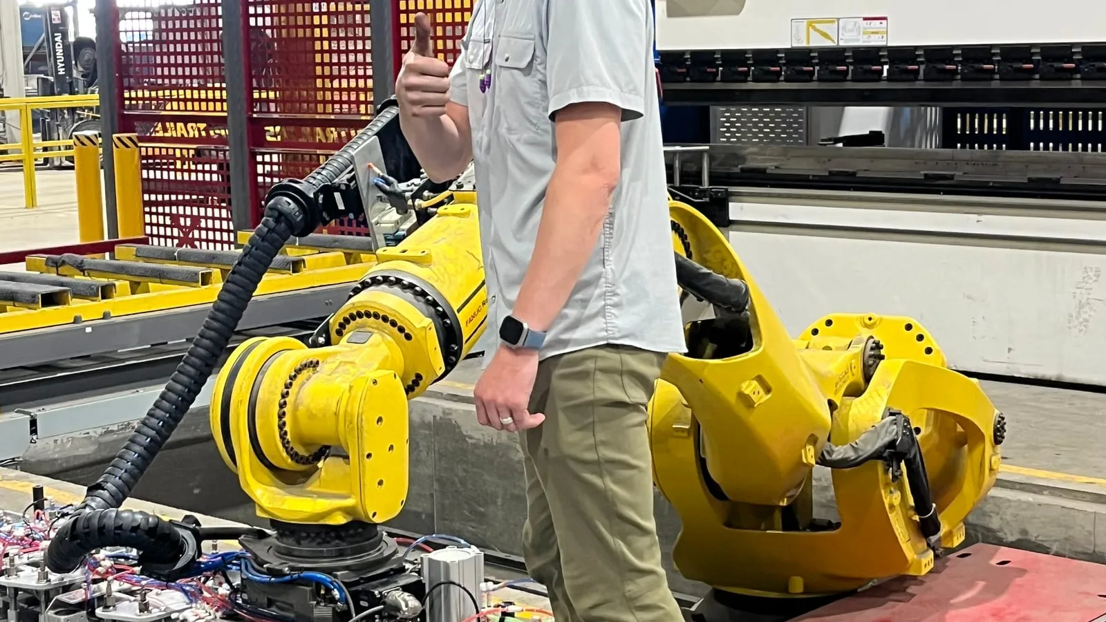 A technician beside a FANUC robot with a long vacuum-cup bar in front of a press brake, on the heavy tandem project