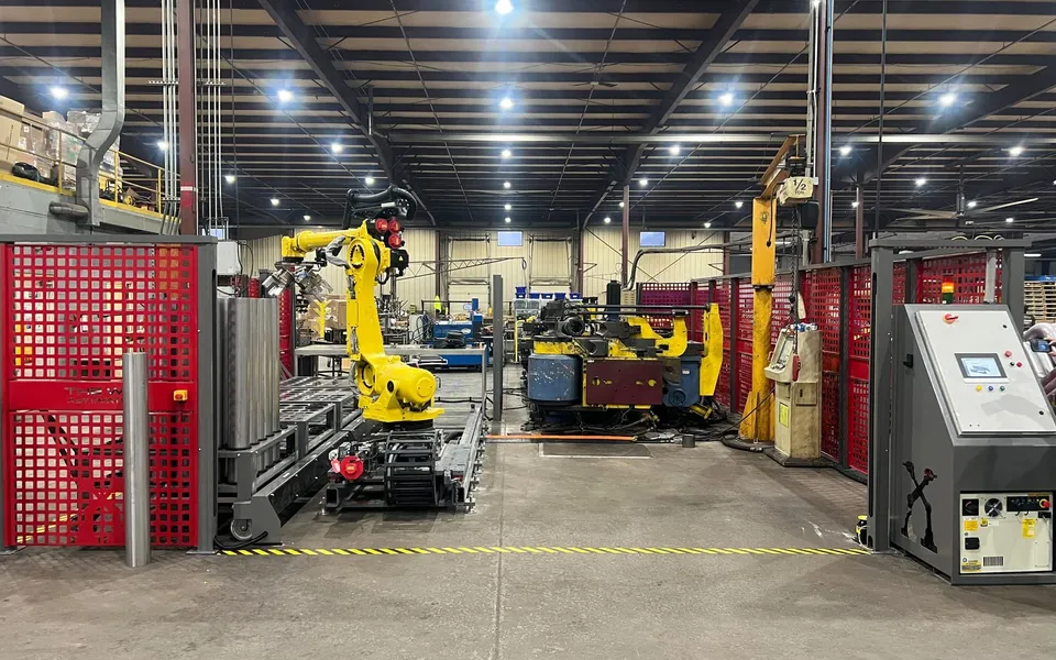 A FANUC robot on a linear rail tending a CNC tube bender inside red safety fencing, control panel at the fence line