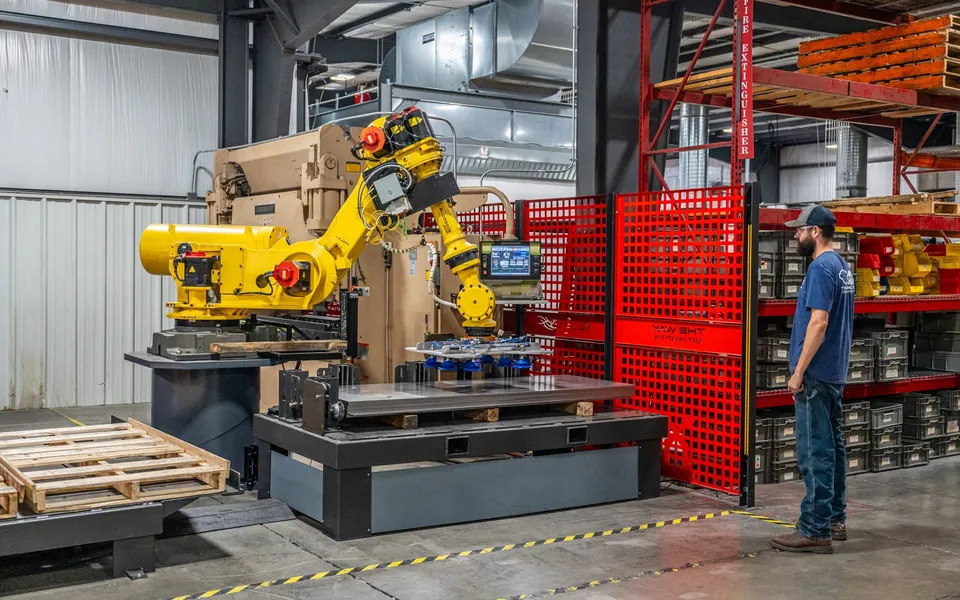 A representative TWA handling cell: a FANUC robot with a vacuum gripper lowering a steel plate onto a pallet station. The customer's floor is not shown.