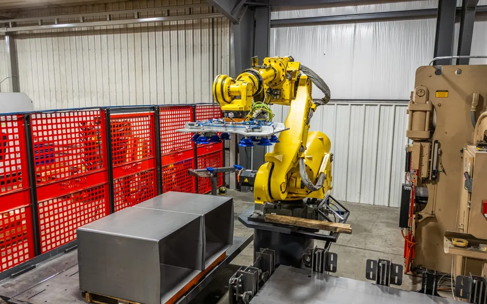 A FANUC robot with a vacuum end effector lifting a sheet at a press brake inside a TWA red-fenced cell
