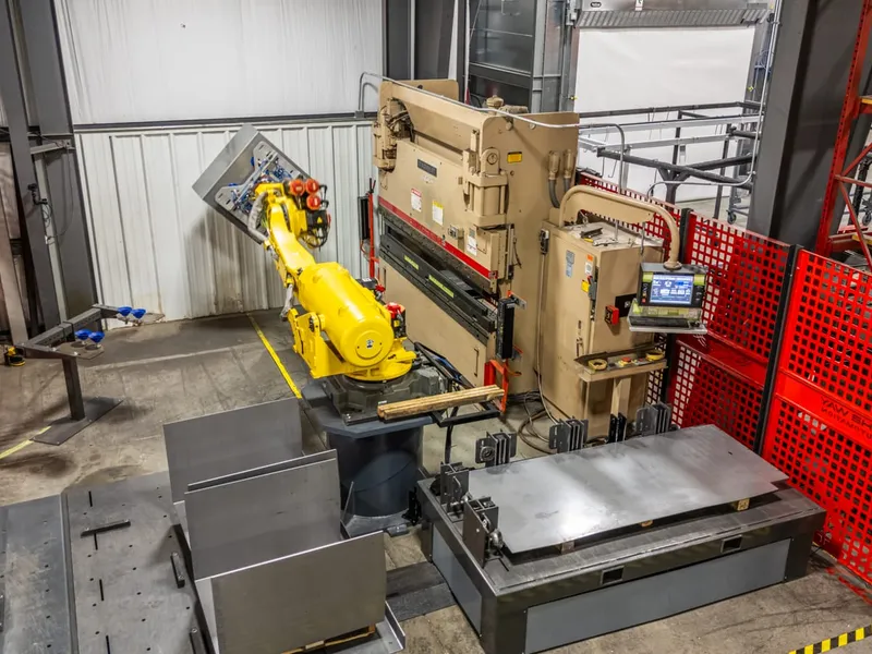 A FANUC robot holding a sheet metal blank in front of a press brake inside a fenced cell
