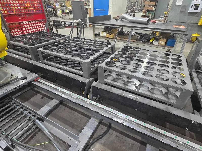 A shop floor cell with part nest trays on racks, a linear rail and a robot base beside a tube bender
