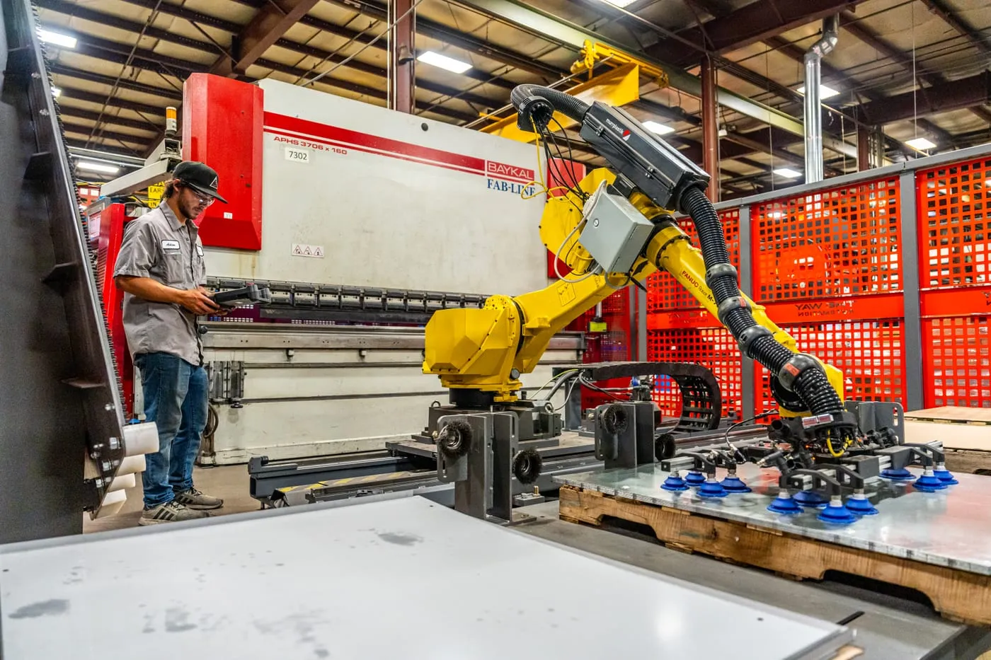 A FANUC robot on a rail loading sheet metal at a Baykal press brake while a TWA technician uses the teach pendant