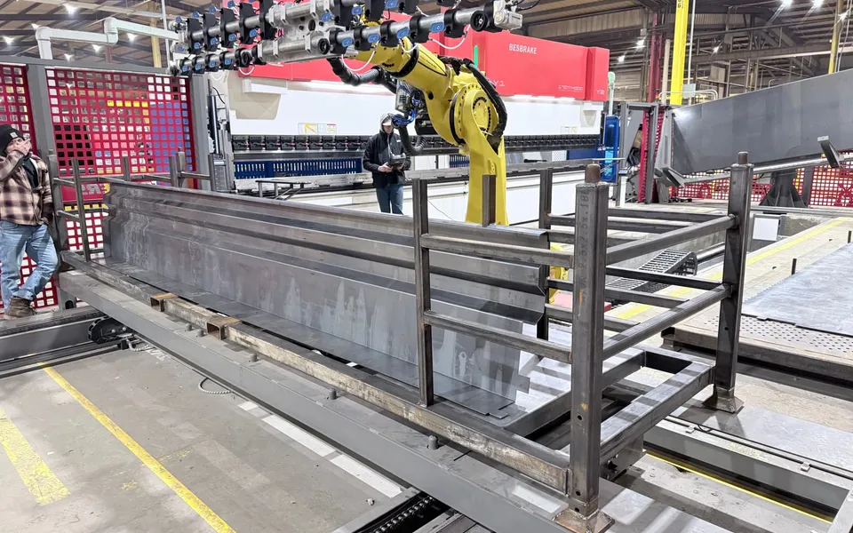 A FANUC robot with a long multi-cup vacuum bar in front of a tandem press brake, a formed trailer panel resting on a steel rack in the foreground