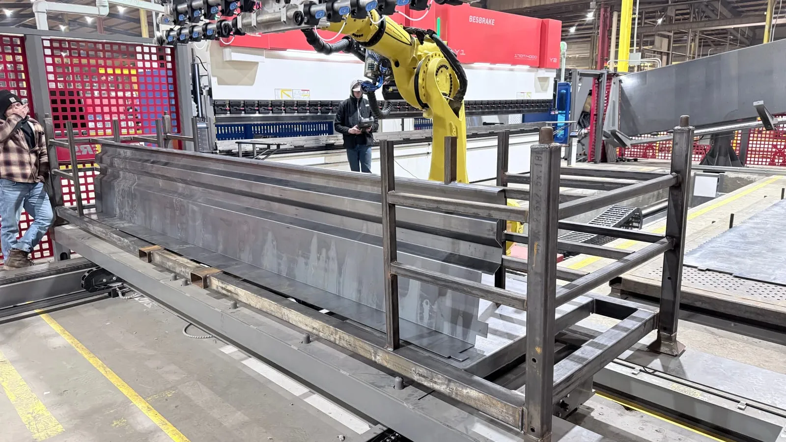 A FANUC robot with a long multi-cup vacuum bar in front of a tandem press brake, a formed trailer panel resting on a steel rack in the foreground