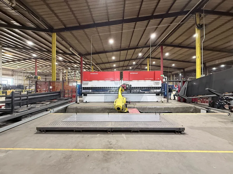 Wide view of the robot rising from its floor pit in front of two press brakes, with a steel blank on the infeed table