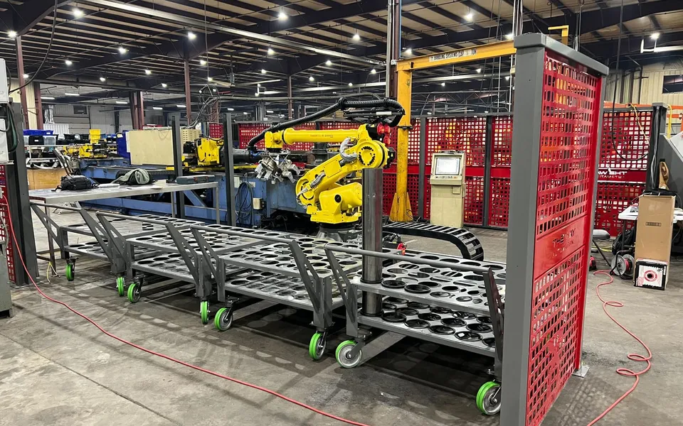 A FANUC robot on a rail tending a tube bender with carts of tube racks inside a fenced cell