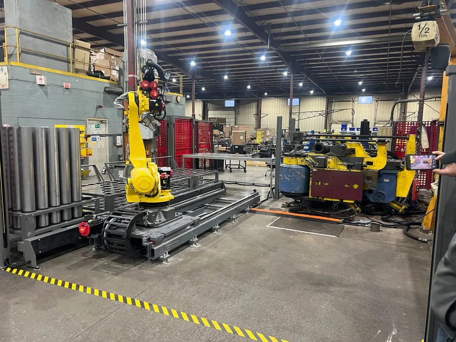The rail-mounted robot between the racks of raw tube on the left and the bender on the right, yellow floor tape marking the cell edge