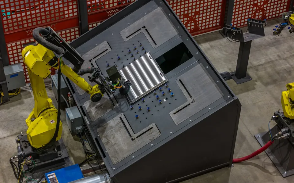 A FANUC robot with a welding torch at a fixture table inside a curtained cell